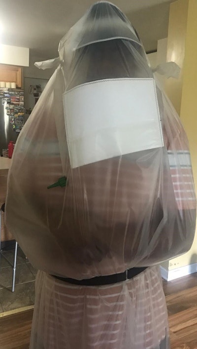 (Above) Disposable isolation device includes an integrated N-95 filter patch; oxygen can be directly connected to the oxygen input nozzle. A hat visor keeps the bag polymer away from face and airway. (Below) An upright patient is assisted by two staff members for careful donning. Images courtesy of Academic Radiology and the National Institutes of Health.