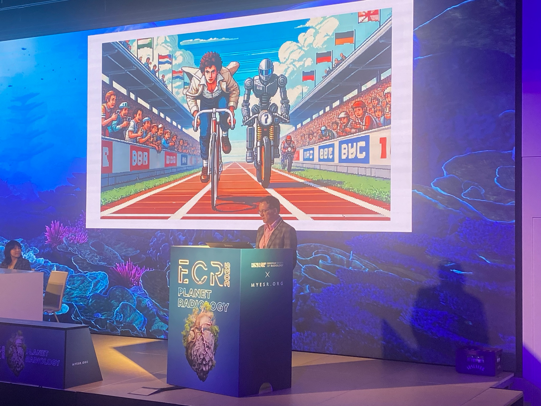 Marc Kohli, MD, discusses current and potential future uses of generative AI in radiology at ECR 2025 in Vienna, Austria. Behind him is an image created by one such generative AI model, which illustrates humans working alongside AI.