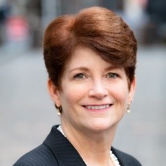 Professional headshot of a smiling woman with auburn hair wearing dark clothing against an urban background