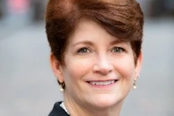 Professional headshot of a smiling woman with auburn hair wearing dark clothing against an urban background