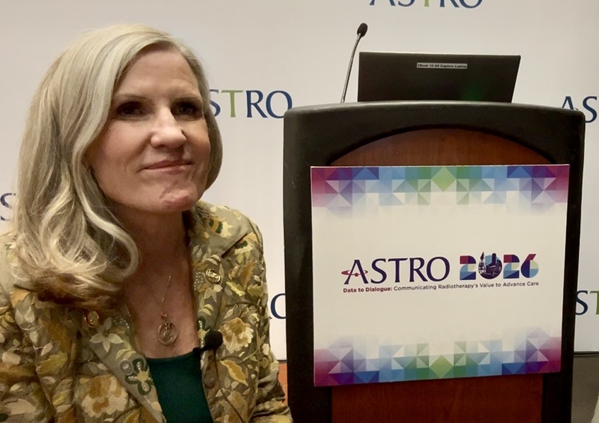 Woman in gold patterned jacket speaking at ASTRO event podium with colorful logo displayed on screen