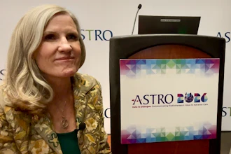 Woman in gold patterned jacket speaking at ASTRO event podium with colorful logo displayed on screen
