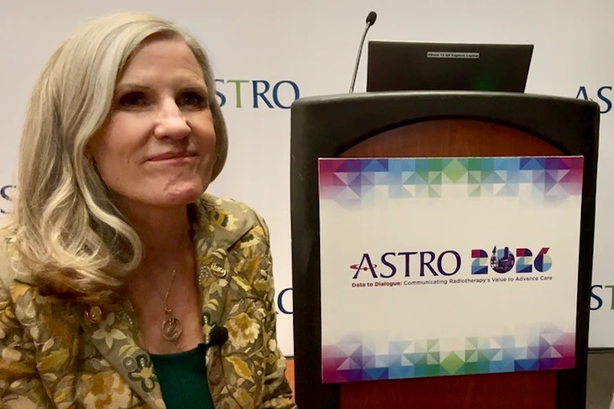 Woman in gold patterned jacket speaking at ASTRO event podium with colorful logo displayed on screen
