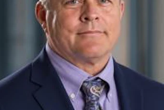 Professional headshot of a middle-aged man wearing a navy blazer and lavender dress shirt with patterned tie