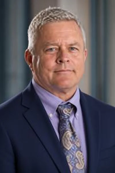 Professional headshot of a middle-aged man wearing a navy blazer and lavender dress shirt with patterned tie