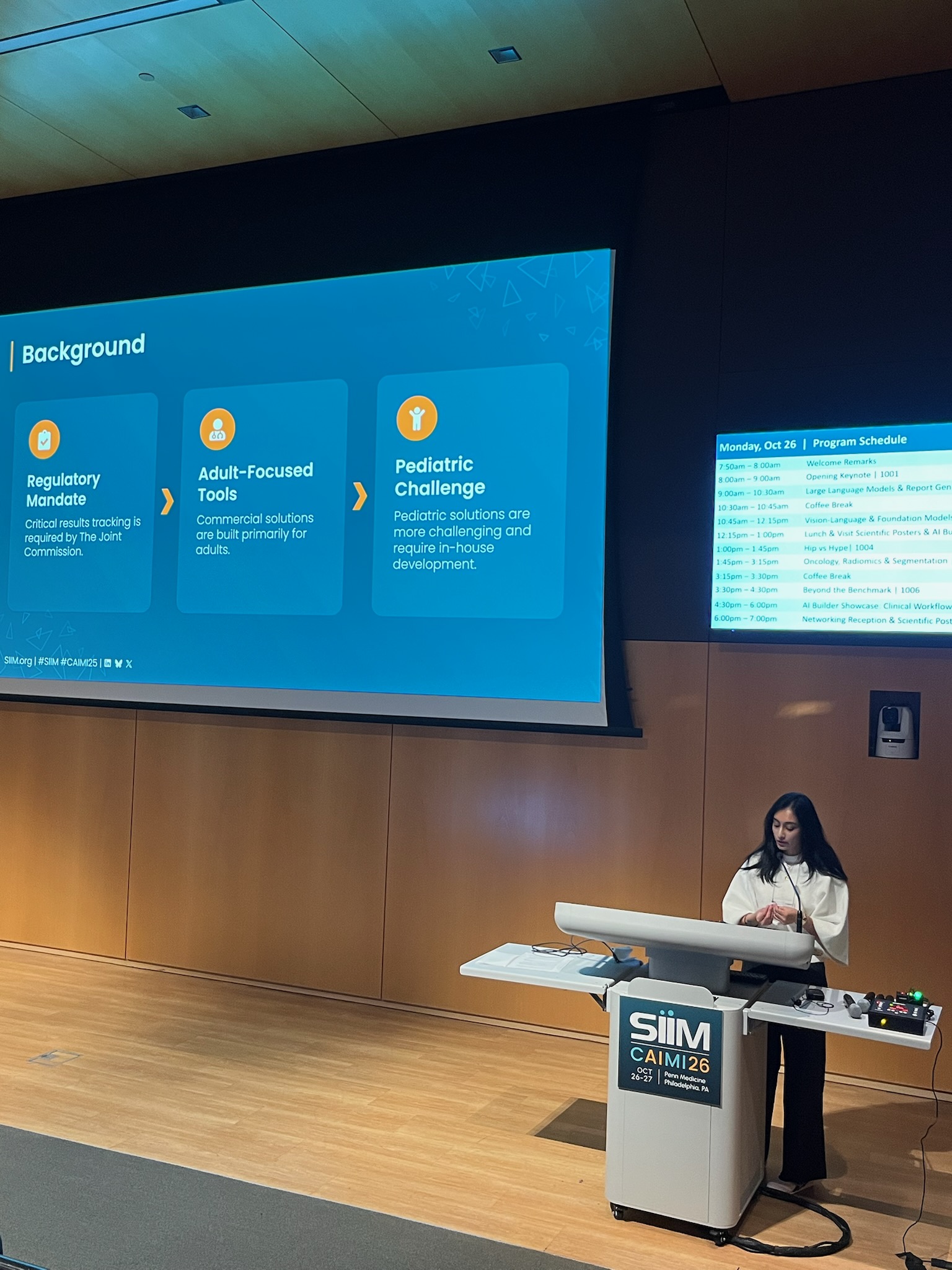 Woman presenting at SIM CAIM26 conference podium with blue program schedule displayed on projection screen