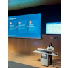 Woman presenting at SIM CAIM26 conference podium with blue program schedule displayed on projection screen