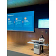 Woman presenting at SIM CAIM26 conference podium with blue program schedule displayed on projection screen