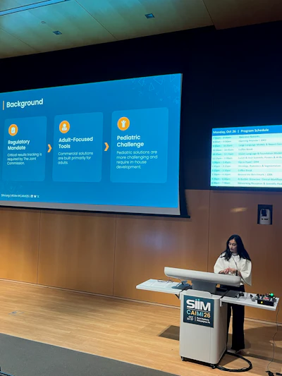 Woman presenting at SIM CAIM26 conference podium with blue program schedule displayed on projection screen