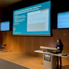 Female presenter delivering conference talk on AI and ICH outcomes at SIM event with projection screens