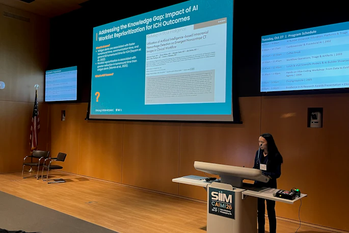 Female presenter delivering conference talk on AI and ICH outcomes at SIM event with projection screens