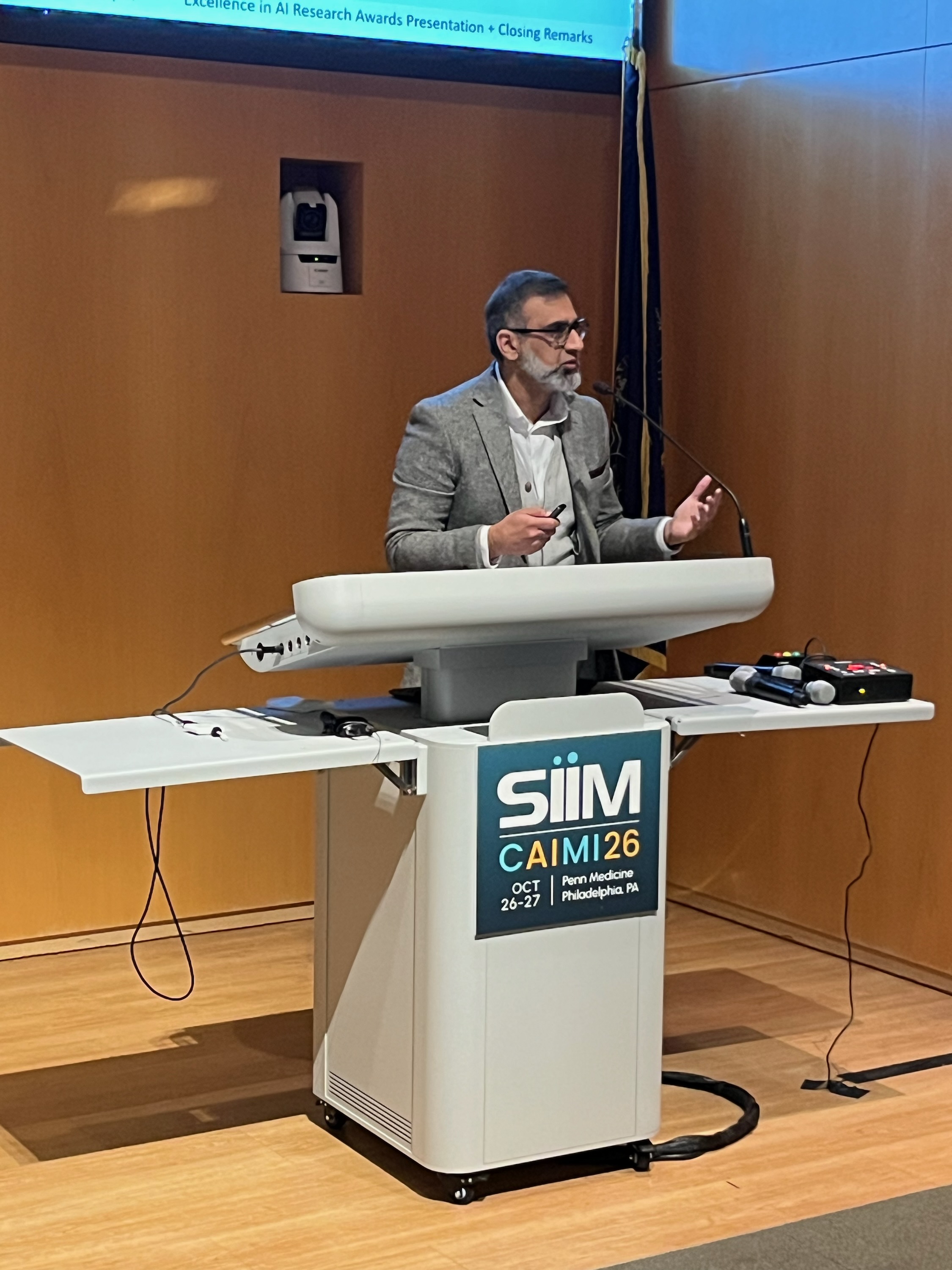 Professional speaker at podium with SIIM CAIMI26 conference logo during AI research awards presentation