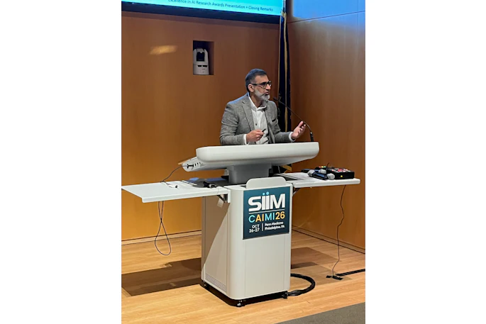 Professional speaker at podium with SIIM CAIMI26 conference logo during AI research awards presentation