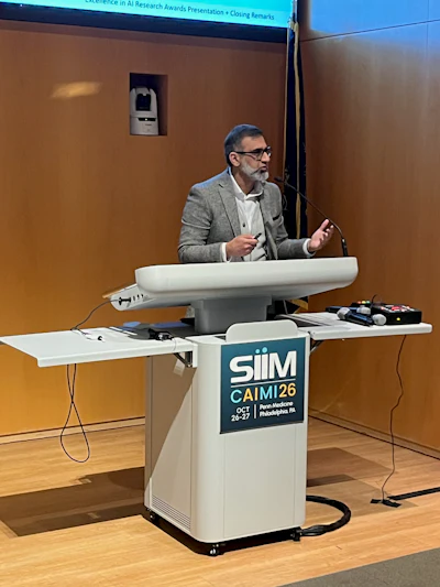 Professional speaker at podium with SIIM CAIMI26 conference logo during AI research awards presentation