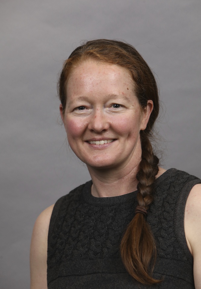 Professional headshot of a woman with reddish-brown hair in a braid, wearing a dark sleeveless top, smiling against a gray background