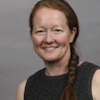 Professional headshot of a woman with reddish-brown hair in a braid, wearing a dark sleeveless top, smiling against a gray background