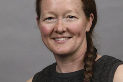Professional headshot of a woman with reddish-brown hair in a braid, wearing a dark sleeveless top, smiling against a gray background