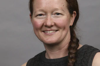 Professional headshot of a woman with reddish-brown hair in a braid, wearing a dark sleeveless top, smiling against a gray background
