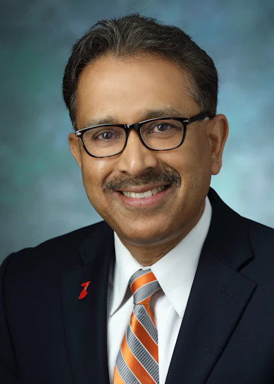 Professional headshot of a man wearing glasses, dark suit, and orange striped tie against blue background