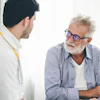 Older man with glasses speaking with younger male healthcare professional in white coat during medical consultation