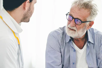 Older man with glasses speaking with younger male healthcare professional in white coat during medical consultation