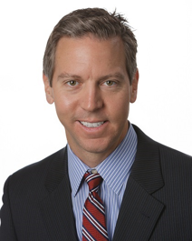 Professional headshot of a man in dark blazer and striped blue shirt with red tie