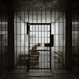 Dimly lit prison cell with barred doors and figure inside, conveying confinement and isolation