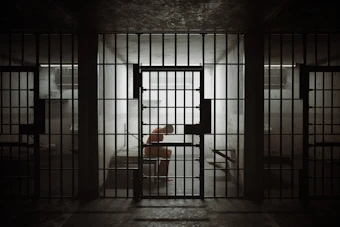 Dimly lit prison cell with barred doors and figure inside, conveying confinement and isolation