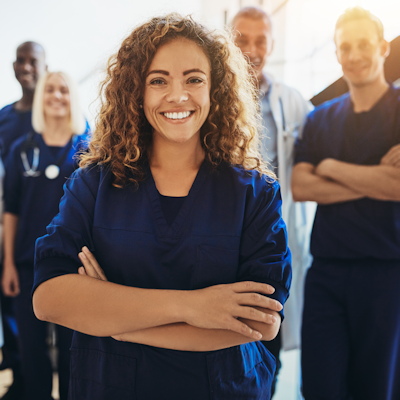 Female healthcare worker in blue scrubs smiling with arms crossed, standing with medical team colleagues in background