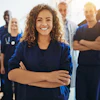 Female healthcare worker in blue scrubs smiling with arms crossed, standing with medical team colleagues in background