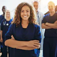 Female healthcare worker in blue scrubs smiling with arms crossed, standing with medical team colleagues in background