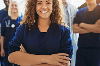Female healthcare worker in blue scrubs smiling with arms crossed, standing with medical team colleagues in background